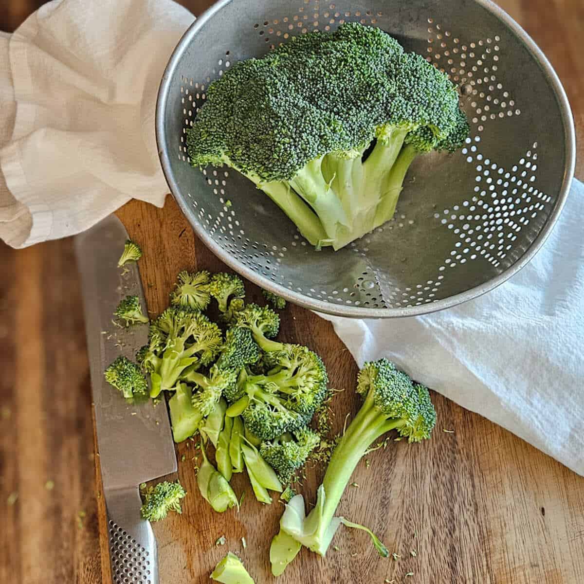 Broccoli in a colander with some on a cutting board being chopped with a knife for the easy Asian broccoli crunch salad.