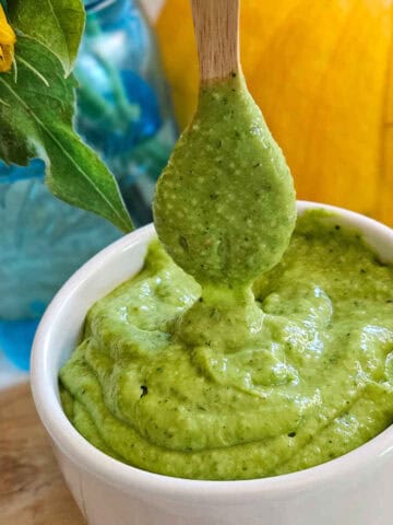 Creamy avocado dressing in a bowl with a spoon being lifted out of the, with flowers in a jar and a pumpkin in the background.
