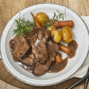 Red wine braised pot roast on a plate with potatoes, carrots, and red wine sauce, garnished with rosemary.
